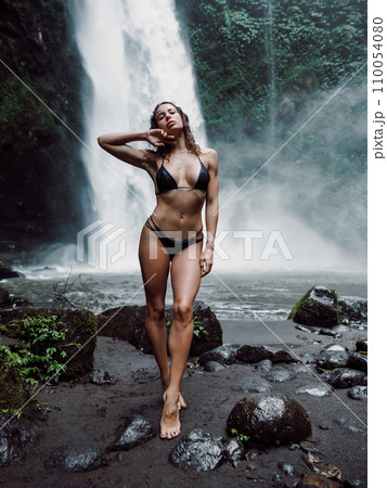 Slim woman in bikini staying near waterfall in Bali. Traveler girl posing on river with powerfull waterfall 110054080