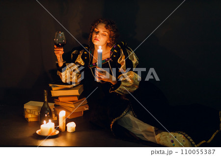 Young adult woman dressed in a medieval dress sitting near ancient books and candle light with bottle of red wine and holding a glass of wine in dark 110055387