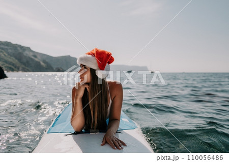 Woman sea sup. Close up portrait of happy young caucasian woman with long hair in Santa hat looking at camera and smiling. Cute woman portrait in a white bikini posing on sup board in the sea 110056486