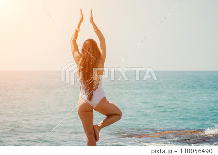 Woman sea yoga. Back view of free calm happy satisfied woman with long hair standing on top rock with yoga position against of sky by the sea. Healthy lifestyle outdoors in nature, fitness concept. 110056490