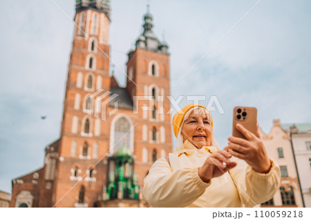 Portrait of 50s old woman with gorgeous smile with teeth and blonde hair against old city center view in Krakow. Takes selfie. 110059218