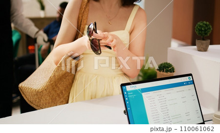 Woman tourist asking for room key card at reception front desk, registering in tropical resort lobby. Traveller dressed in summer clothes having reservation for holiday trip. Handheld shot. Close up. Woman tourist asking for room key card at reception front desk, registering in tropical resort lobby. Traveller dressed in summer clothes having reservation for holiday trip. Handheld shot. Close up. 110061062