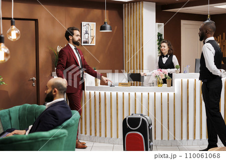 Front desk staff greets businessman with suitcase baggage, travelling on work trip and staying at luxury hotel. Formal person entering lobby to ask about room reservation, registration. Front desk staff greets businessman with suitcase baggage, travelling on work trip and staying at luxury hotel. Formal person entering lobby to ask about room reservation, registration. 110061068