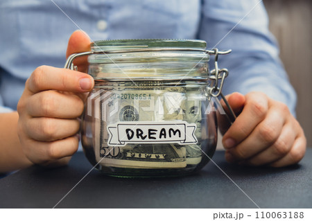 Female hands holding Glass jar full of American currency dollars cash banknote with text DREAM. Preparation saving money. Moderate consumption and economy Female hands holding Glass jar full of American currency dollars cash banknote with text DREAM. Preparation saving money. Moderate consumption and economy 110063188