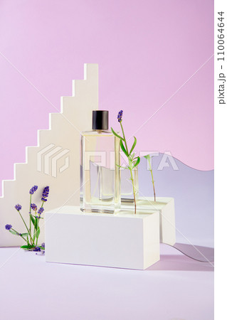 A perfume bottle on a white podium, mirrored reflection, and lavender blooms against a light purple backdrop create an enticing mockup for perfume advertising. 110064644