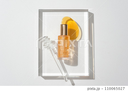A serum bottle with yellow essence particles, honey drops and a honey drizzle on a glass podium on a white background. Cosmetic mockup for advertising. 110065027