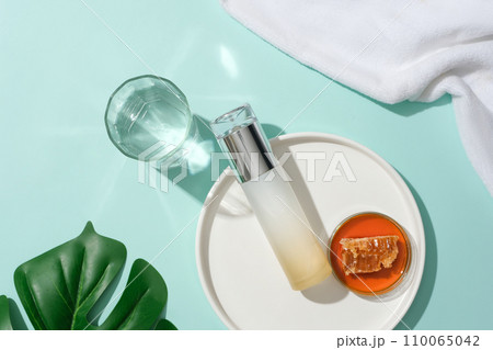 Honey and an unlabeled cosmetic bottle are displayed on a white ceramic plate, a glass of water, green leaves and a towel on a blue background. View from above. 110065042