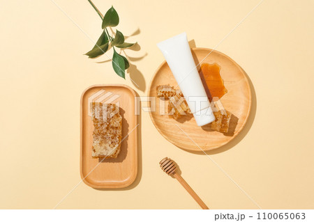 Beeswax is stored on a wooden tray, an unlabeled cosmetic tube and honey on a round wooden plate, a honey drizzle and green leaves on a pastel background. Ideal space for cosmetics display. Beeswax is stored on a wooden tray, an unlabeled cosmetic tube and honey on a round wooden plate, a honey drizzle and green leaves on a pastel background. Ideal space for cosmetics display. 110065063