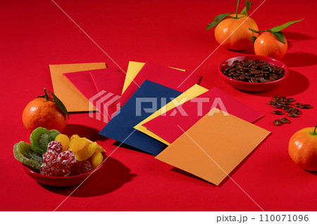 Colorful lucky money envelopes, a plate of dried fruit, a plate of melon seeds and tangerines are placed on a red background. Festive atmosphere, New Year. 110071096