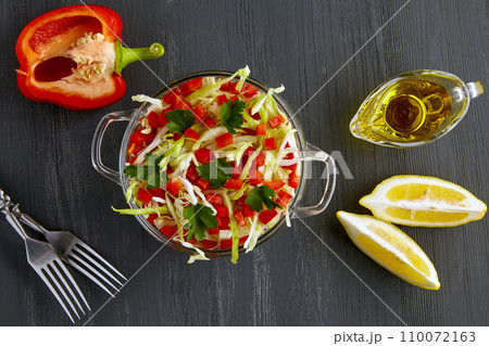 Fresh vegetable salad of cabbage and sweet peppers in a glass bowl on a black wooden background surrounded by ingredients 110072163