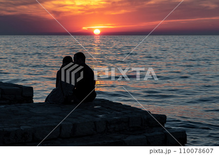 Couple Of Young Adults Sit At The Beach Of The Mediterranean Sea And Looks At The Sunset Near The City Of Porec In Croatia Couple Of Young Adults Sit At The Beach Of The Mediterranean Sea And Looks At The Sunset Near The City Of Porec In Croatia 110078607