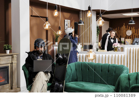 Male guest in snow gear enjoys comfort of lounge area while planning winter holiday activities with personal computer. Tourists in ski hotel reception with wintersports equipment and digital device. Male guest in snow gear enjoys comfort of lounge area while planning winter holiday activities with personal computer. Tourists in ski hotel reception with wintersports equipment and digital device. 110081389