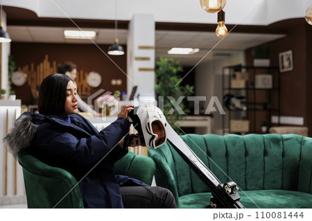 Asian woman relaxes in hotel lobby and prepares her snow gear for wintersports activity. Female tourist with winter jacket grasping her ski goggles skiings skis and helmet in winter mountain resort. 110081444