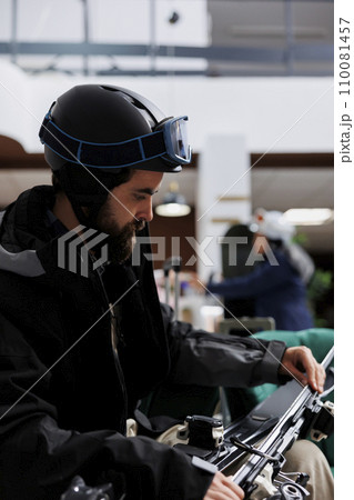Close-up of caucasian traveler adjusting ski gear in hotel lobby. Male tourist, wearing winter jacket and ski goggles, ensuring safety of wintersports equipment. Ready to explore ski resort for skiing 110081457