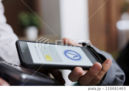 Close-up shot of tourist making contactless payment via mobile phone using NFC technology. Detailed view of pair of hands grasping pos terminal and smartphone, paying winter holiday reservation. 110081463