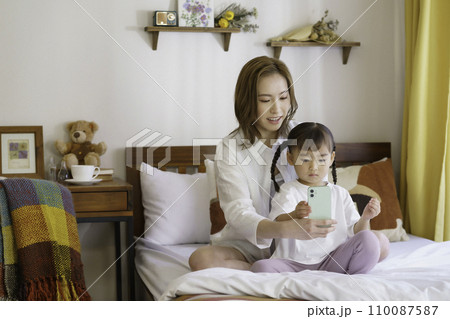 ベッドの上でスマホを操作する茶髪の若いママと4歳の女の子 ベッドの上でスマホを操作する茶髪の若いママと4歳の女の子 110087587