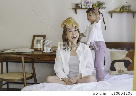 ベッドの上でクマのおぬいぐるみをママの頭に乗せて飛び跳ねる4歳の女の子 110091206