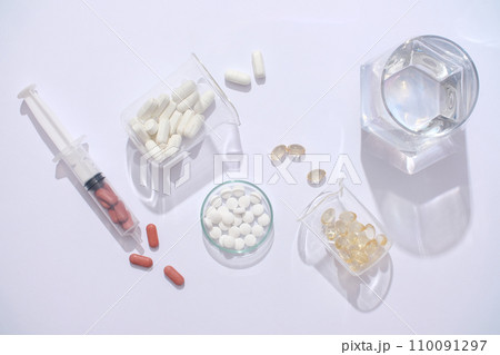 On a white backdrop, a glass of water, syringe, and medicines arranged on a transparent petri dish and lab beaker. Essential aids in supporting the treatment process. On a white backdrop, a glass of water, syringe, and medicines arranged on a transparent petri dish and lab beaker. Essential aids in supporting the treatment process. 110091297