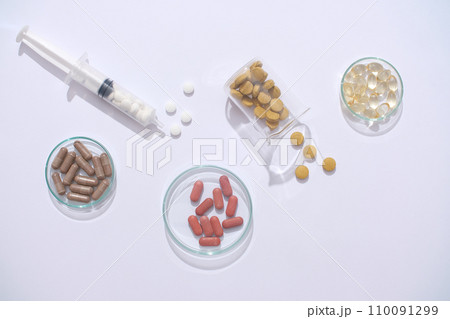 Against a minimalist backdrop, a glass of water, syringe, and medicines are showcased on a transparent petri dish and beaker. Medicine play a crucial role in supporting the treatment process. Against a minimalist backdrop, a glass of water, syringe, and medicines are showcased on a transparent petri dish and beaker. Medicine play a crucial role in supporting the treatment process. 110091299