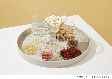Close-up of a glass of water and a variety of dietary supplements displayed in glassware on a ceramic plate. Dietary supplements are produced in the form of tablets, capsules, liquids, etc. 110091315