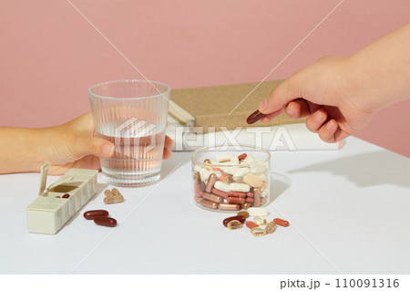One hand is holding a glass of water, another hand is holding a pill taken from a glass medicine jar. On the table there is a notebook and a weekly pill organizer. Medicine theme. One hand is holding a glass of water, another hand is holding a pill taken from a glass medicine jar. On the table there is a notebook and a weekly pill organizer. Medicine theme. 110091316