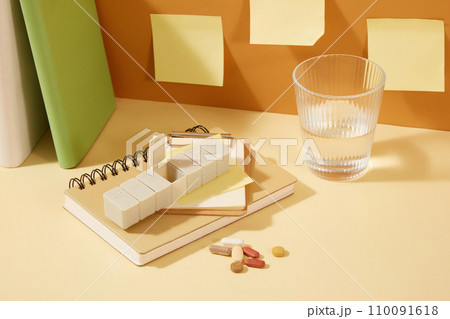 Close-up of a corner of a desk with notebooks, notes, a box of daily pills and a glass of water. Space for advertising with a medicine theme. Close-up of a corner of a desk with notebooks, notes, a box of daily pills and a glass of water. Space for advertising with a medicine theme. 110091618