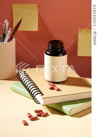 An unlabeled bottle of dietary supplements is placed on top of two notebooks. Yellow sticky notes are stuck on the wall. Mockup for medicine advertising. 110091619