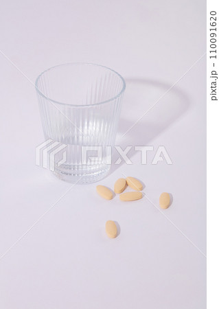 A glass of water and pastel pink pills are displayed on a white background. Minimalist space. Advertising drugs and functional foods. Commercial images. 110091620