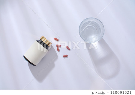 Top view of an unbranded medicine bottle, pills and a glass of water on a white background with drop shadow. Blank label for lettering design. New drug launch. Top view of an unbranded medicine bottle, pills and a glass of water on a white background with drop shadow. Blank label for lettering design. New drug launch. 110091621