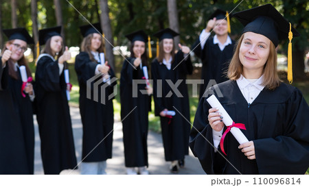 Group of happy students in graduation gowns outdoors. A young girl with a diploma in her hands in the foreground. 110096814