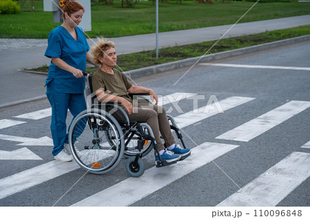 Red-haired nurse pushing an elderly woman in a wheelchair across the road. Red-haired nurse pushing an elderly woman in a wheelchair across the road. 110096848