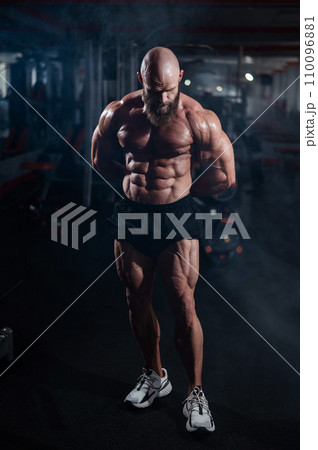 Muscular bald man posing in shorts. Bodybuilder showing off his shape in the gym.  110096881