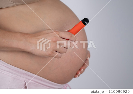 A pregnant woman smokes a vape. A girl holds an electronic cigarette against the background of her bare tummy. Copy space.  110096929