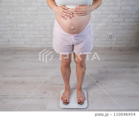 Top view of a pregnant woman in home clothes standing on an electronic scale and holding her hands on her tummy.  110096954