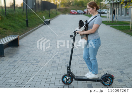 A woman rides an electric scooter with a dog in a backpack. Pappilion Spaniel Continental in a sling. 110097267