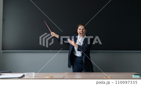 Red-haired caucasian woman in a trouser suit. Angry female teacher with a pointer screaming at the blackboard.  110097335
