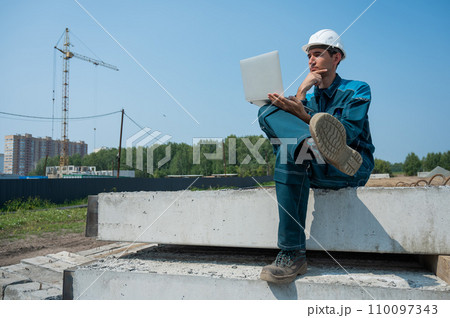 Caucasian male builder in hardhat sits on floor slabs and uses laptop at construction site.  110097343