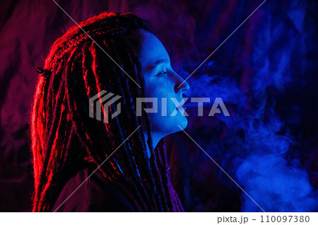 Profile of caucasian girl with dreadlocks smokes a vape in red blue light.  110097380