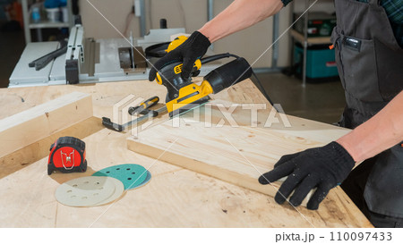 A man using an orbital wood sander in a workshop. Close-up of a carpenter's hands. A man using an orbital wood sander in a workshop. Close-up of a carpenter's hands. 110097433