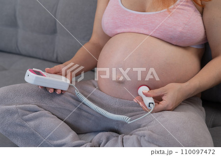 A pregnant woman uses a home doppler while sitting on the couch. Close-up of the abdomen. 110097472