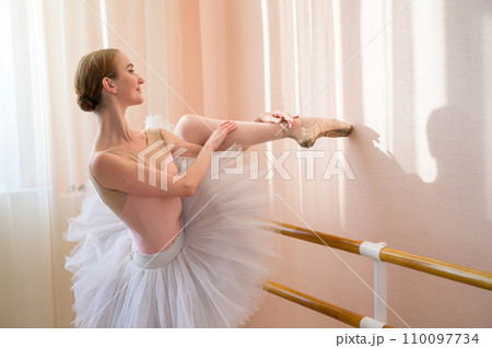 Beautiful graceful ballerina is practicing in the hall against the background of a window. Slender woman in tutu and pointe shoes in a dance class. Beautiful graceful ballerina is practicing in the hall against the background of a window. Slender woman in tutu and pointe shoes in a dance class. 110097734