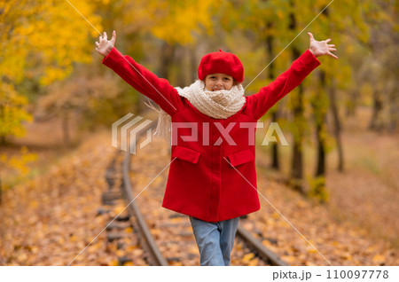 Caucasian girl in a red coat and beret walks along the railway tracks in the park in autumn. Caucasian girl in a red coat and beret walks along the railway tracks in the park in autumn. 110097778