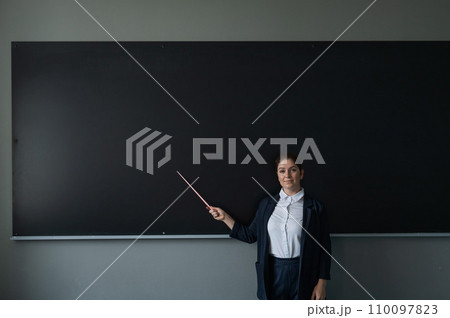 Red-haired caucasian woman in a trouser suit. Smiling female teacher with a pointer at the blackboard.  110097823