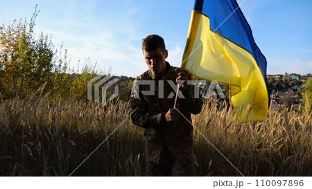 Young male military in uniform walks with waving flag of Ukraine at countryside. Soldier of ukrainian army going with lifting national banner outdoor. Victory against russian aggression concept Young male military in uniform walks with waving flag of Ukraine at countryside. Soldier of ukrainian army going with lifting national banner outdoor. Victory against russian aggression concept 110097896