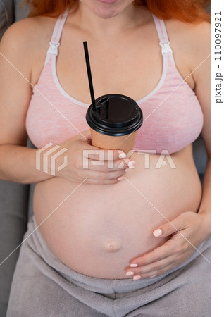Faceless pregnant woman in 3rd trimester holding a coffee glass on her belly.  110097921