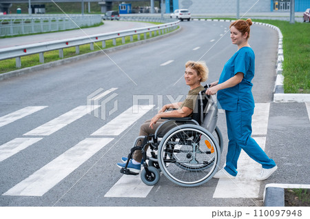 Red-haired nurse pushing an elderly woman in a wheelchair across the road.  110097948