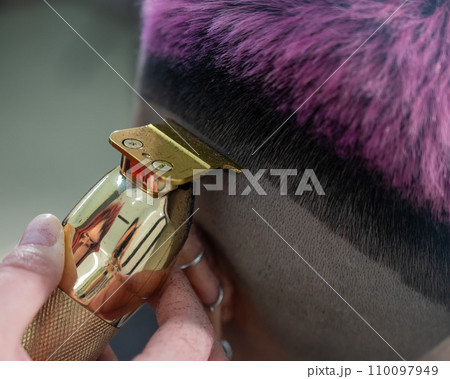 The hairdresser shaves the temple of a female client. Rear view of a woman with short pink hair in a barbershop. The hairdresser shaves the temple of a female client. Rear view of a woman with short pink hair in a barbershop. 110097949