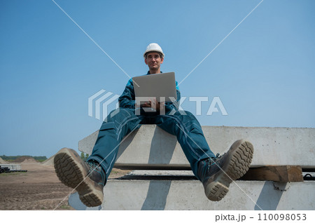 Caucasian male builder in hardhat sits on floor slabs and uses laptop at construction site. Caucasian male builder in hardhat sits on floor slabs and uses laptop at construction site. 110098053