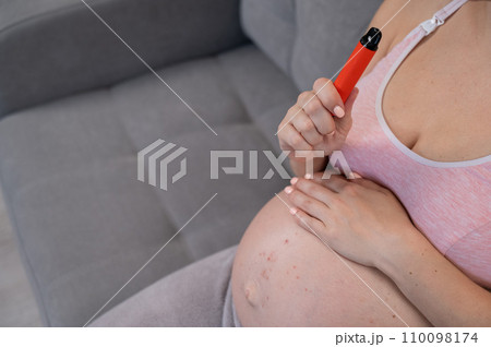 A pregnant woman smokes a vape while sitting on the couch. Close-up of the belly with allergies. A pregnant woman smokes a vape while sitting on the couch. Close-up of the belly with allergies. 110098174