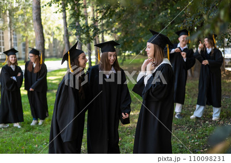 A group of graduates in robes congratulate each other on their graduation outdoors. A group of graduates in robes congratulate each other on their graduation outdoors. 110098231